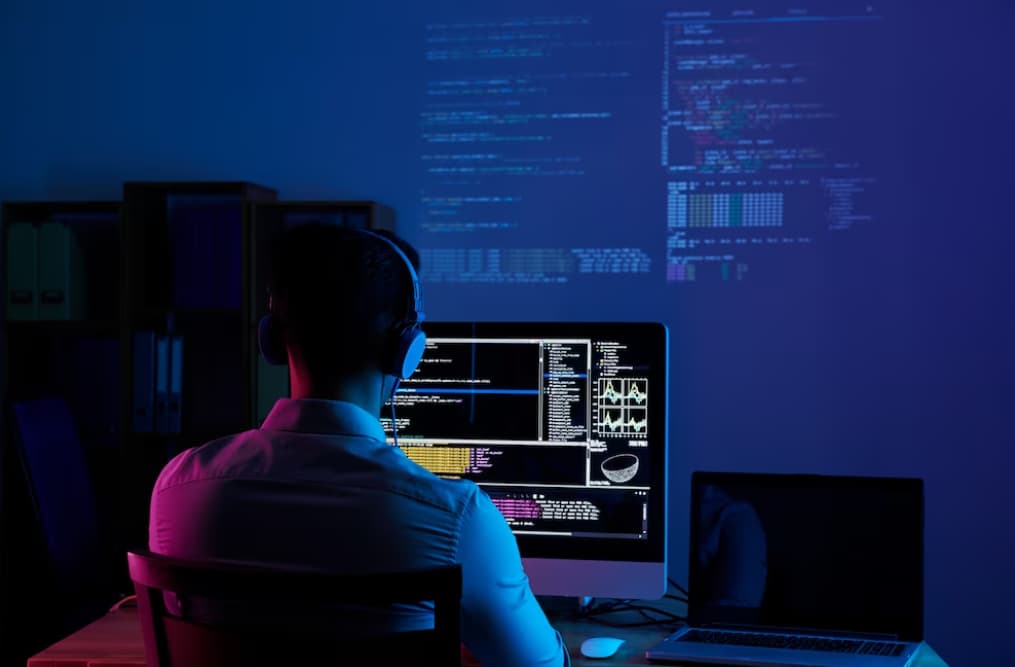 A programmer works on code in a dark room with multiple screens