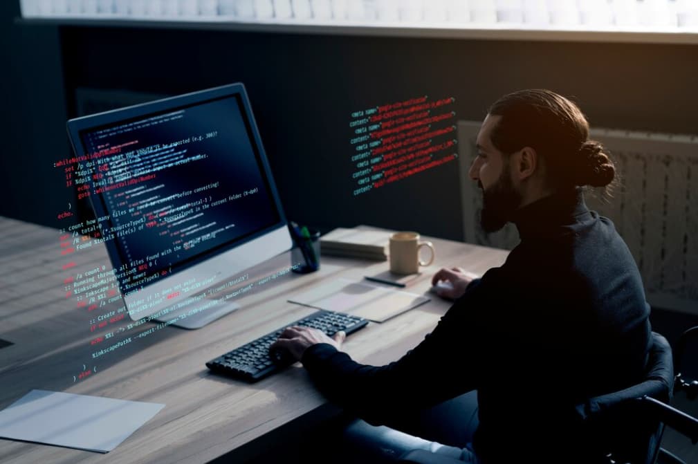 A man with a bun coding on a computer in a dimly lit office